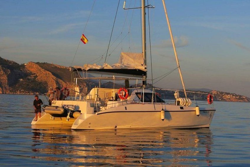 A sailboat with a flag, calm water, and rocky hills in the background. 1