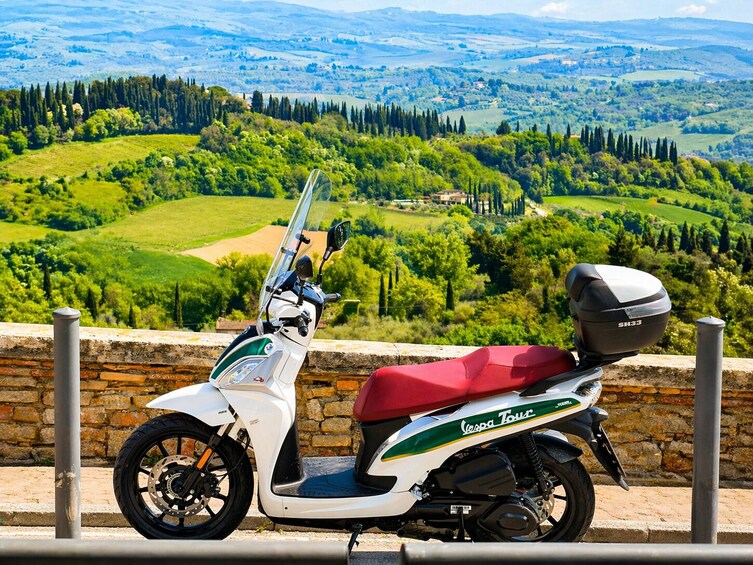 Valkoinen Vespa Tour -skootteri, jossa on punainen istuin, on pysäköity kivimuurille, jolta avautuu näkymä kauniille maisemalle. 4