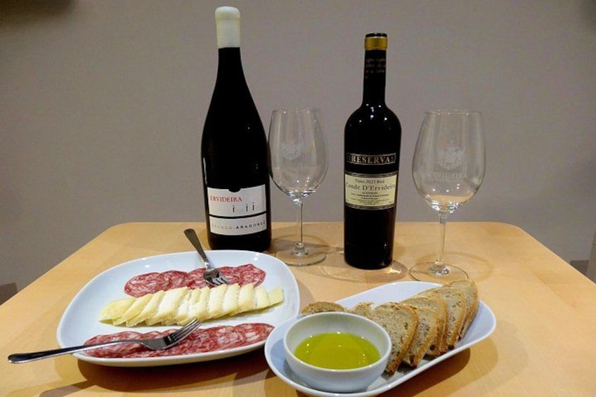 Una mesa con una botella de vino, dos copas, un plato con salami en rodajas, un plato con pan y un bol de aceite de oliva. 5