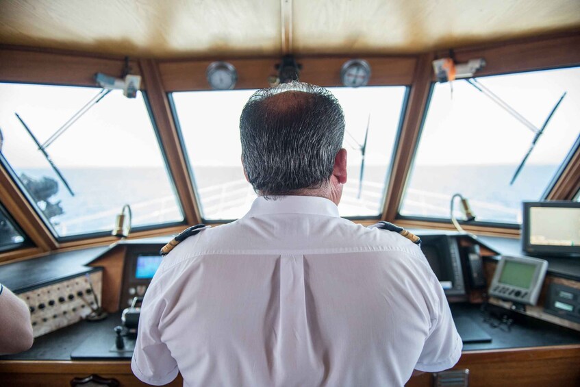 A man in a white shirt from behind, looking out of a ship's bridge with multiple control panels and windows. 4