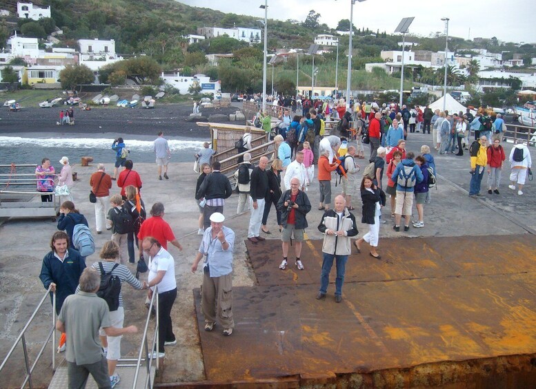 A crowded pier with people walking and standing, overlooking a beach with boats and buildings in the background. 5