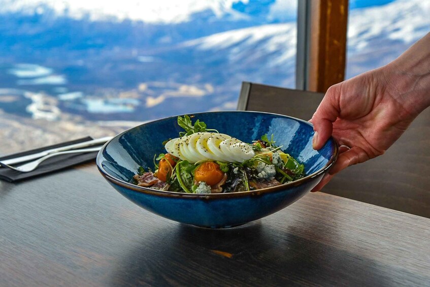 A hand holding a blue bowl of salad with a mountainous landscape in the background. 3