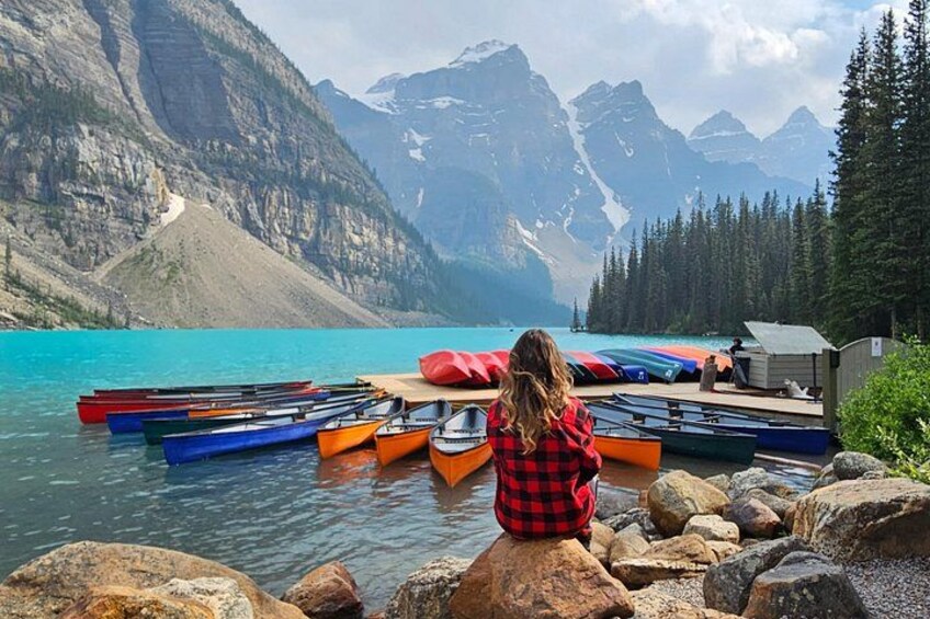 A person sitting on rocks by a lake with canoes, surrounded by mountains and trees. 2