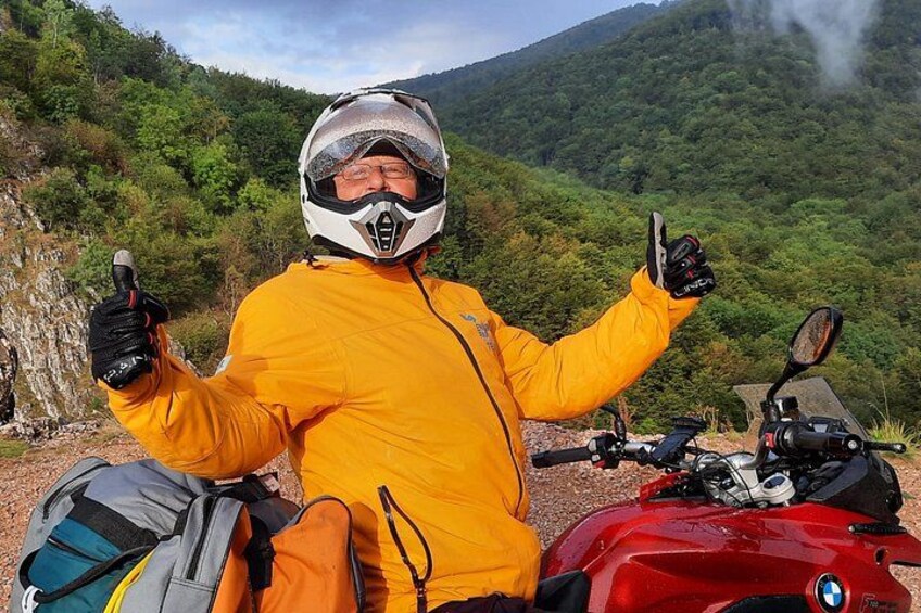 A person in a yellow jacket and helmet sitting on a red motorcycle with a backpack beside them, surrounded by a mountainous landscape. 2