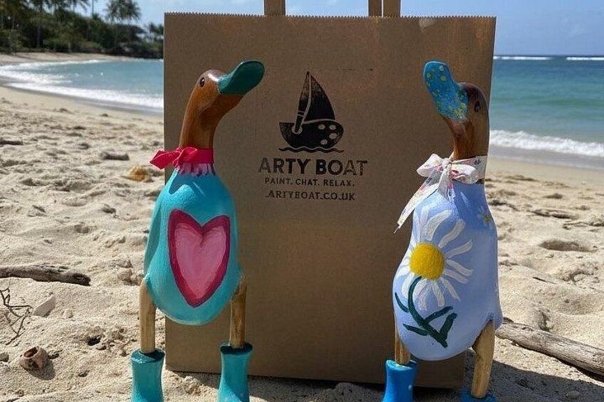Two painted wooden figures on a beach with a cardboard box labeled 'ARTY BOAT' in the background. 2