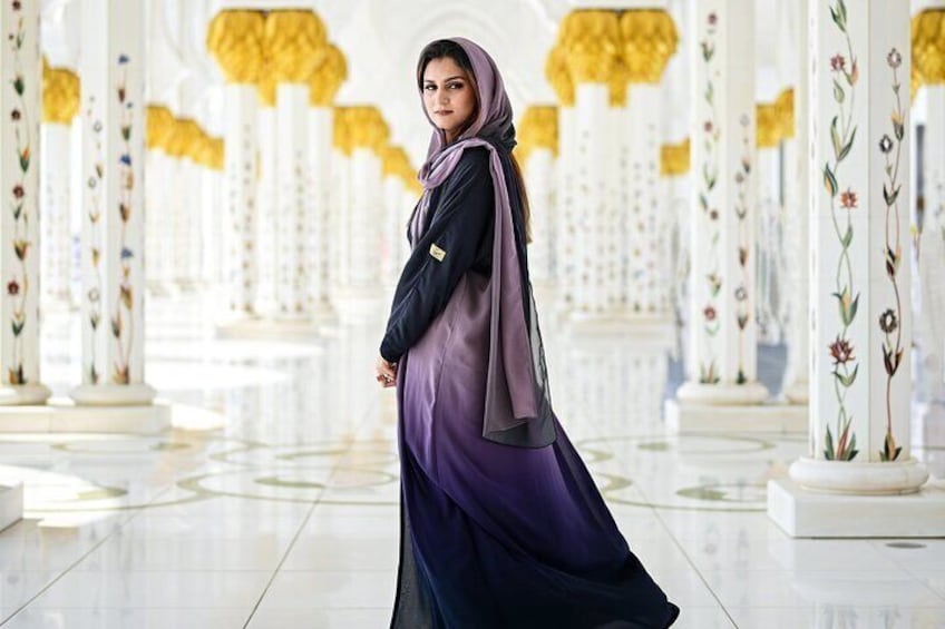 Una persona con un vestido largo morado y un pañuelo en la cabeza caminando por un interior amplio y ricamente decorado con columnas blancas y diseños florales. 3