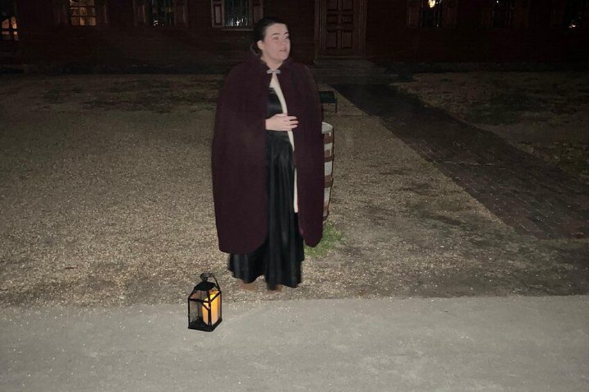 A person in a dark dress and cape standing on a concrete surface with a lantern beside them. 1