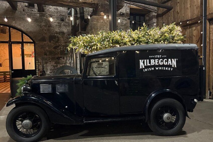 A vintage black truck with 'Kilbeggan Irish Whiskey' written on the side, parked indoors with a rustic interior background. 4
