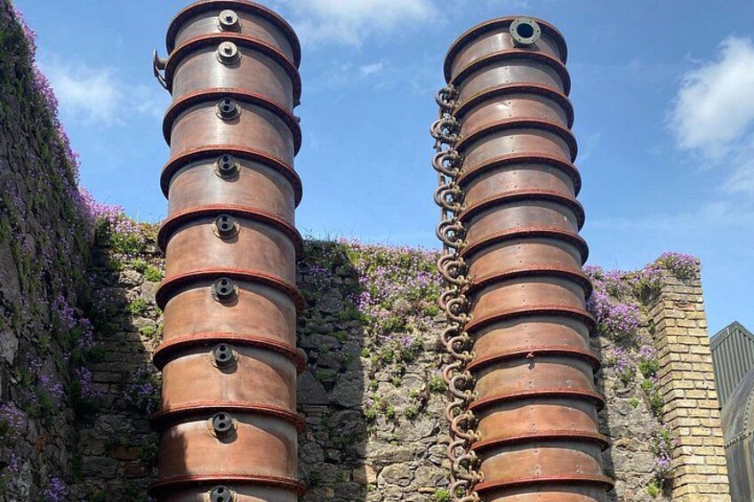 Two cylindrical metal structures with rivets and chains, set against a stone wall with purple flowers. 3