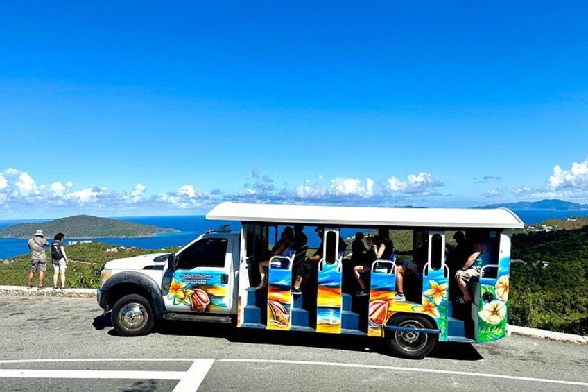 A colorful bus with passengers, parked on a road with a scenic view of the ocean and hills in the background. 3