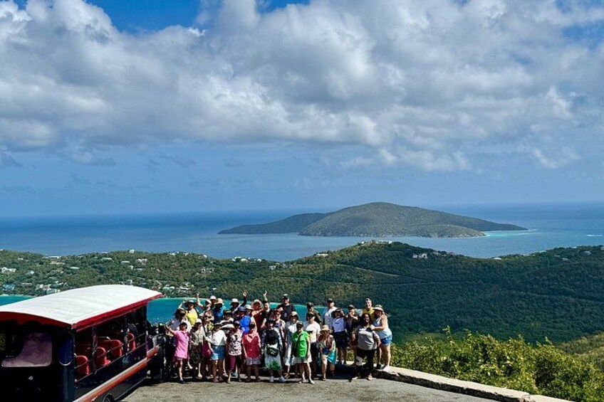 A group of people on a road with a bus, overlooking a coastal landscape with a mountain and the sea. 2