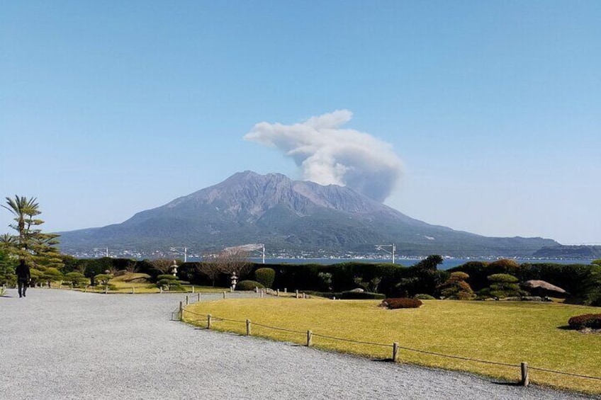 A mountain with a smoke plume, a paved path, and a fenced grassy area with shrubs. 1
