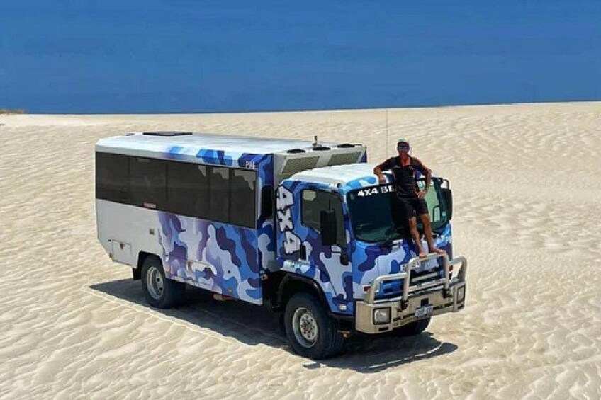A camouflaged bus parked on a sandy beach with a person standing on the driver's side. 3