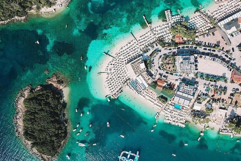 Vista aérea de un complejo turístico costero con una playa en forma de media luna, aguas turquesas cristalinas y una pequeña isla cerca. 1