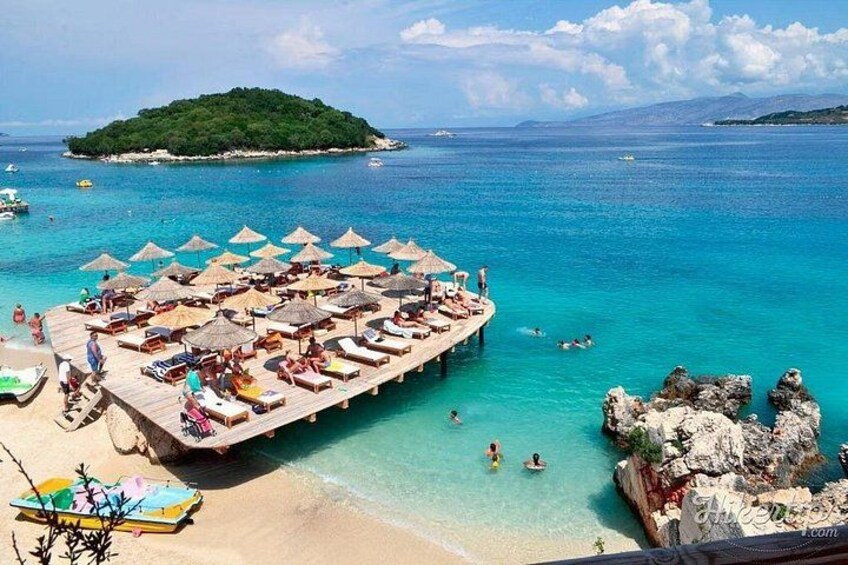 Una playa con un muelle de madera, sillas de descanso y sombrillas. Se ven personas nadando y tomando el sol. 2