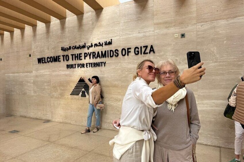 Eine Gruppe von Personen macht ein Selfie vor einer Wand mit dem Text "Welcome to the Pyramids of Giza" und einem Pyramiden-Symbol. 1