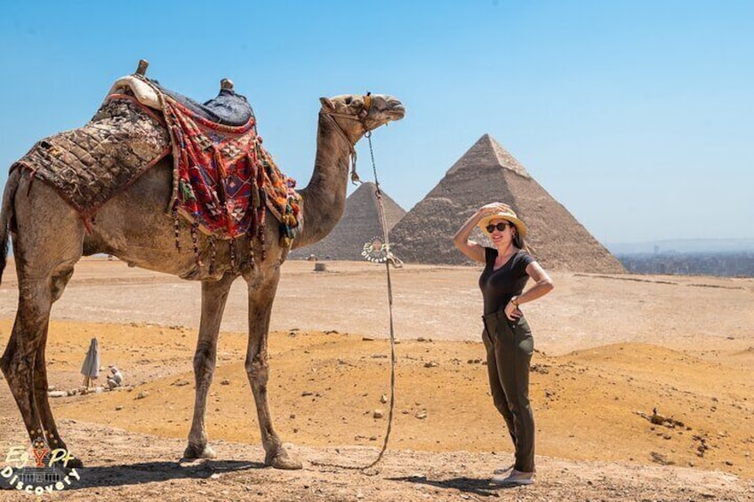 Ein Kamel mit Sattel und eine Person vor einer Pyramide. 3