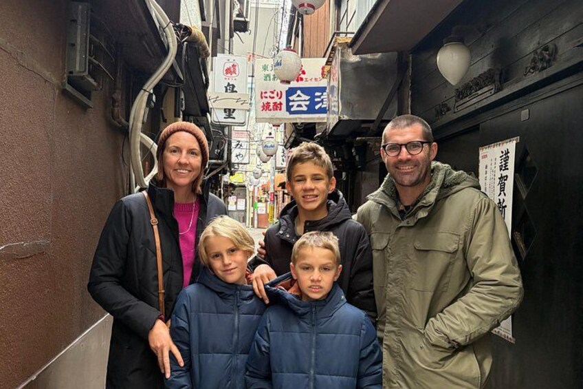Un callejón estrecho con gente de pie. Hay letreros con caracteres japoneses y una puerta negra con un cartel encima. 5