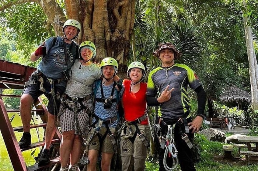 A group of people with helmets and harnesses, standing near a tree and a wooden structure. 3