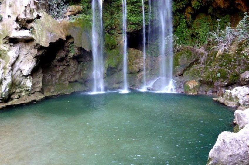 Ein natürlicher Wasserfall stürzt in einen klaren Teich, umgeben von felsigem Gelände und Grünpflanzen. 1