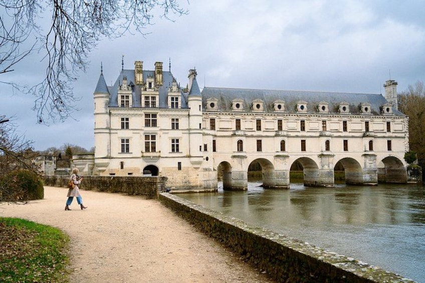 A historic building with a river in front, a person walking on the path, and a bridge. 4