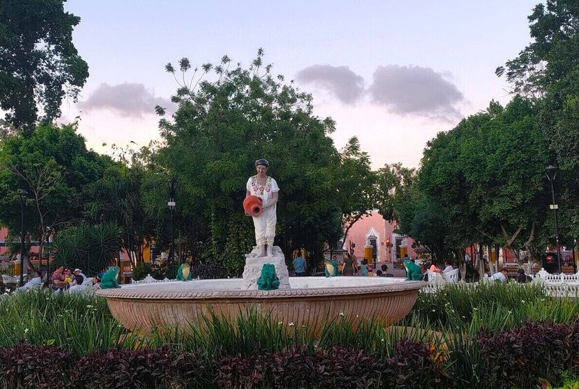 A statue of a figure holding a pot, surrounded by a circular fountain, with trees and people in the background. 4