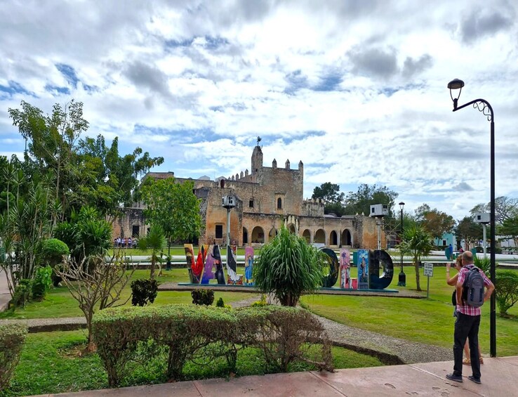 A historic building with a tower, surrounded by a well-maintained garden with various plants and a sculpture of a person. 2