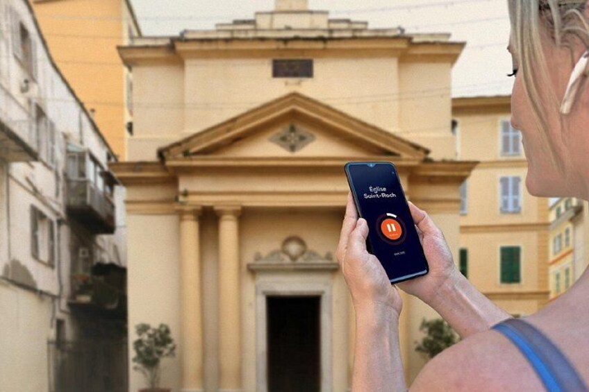 Una persona sosteniendo un teléfono inteligente con la interfaz de una aplicación de música, de pie frente a un edificio de arquitectura clásica. 5