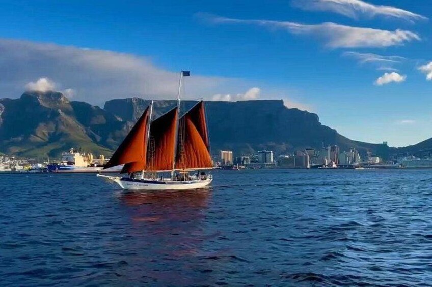 Een zeilboot met rode zeilen op rustig water, een stadsachtergrond en bergen op de achtergrond. 1