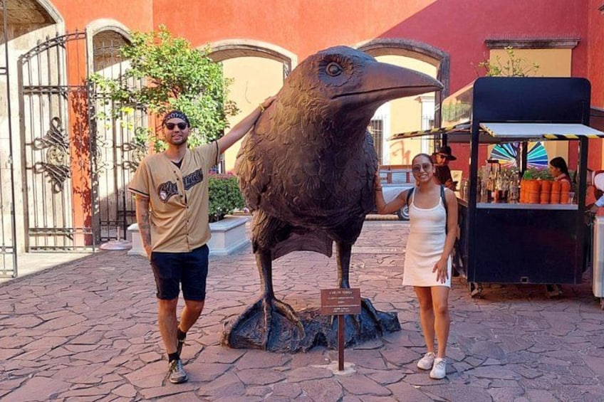 Een man en een vrouw poseren met een groot vogelbeeld in een openluchtsetting, met een kiosk op de achtergrond. 3