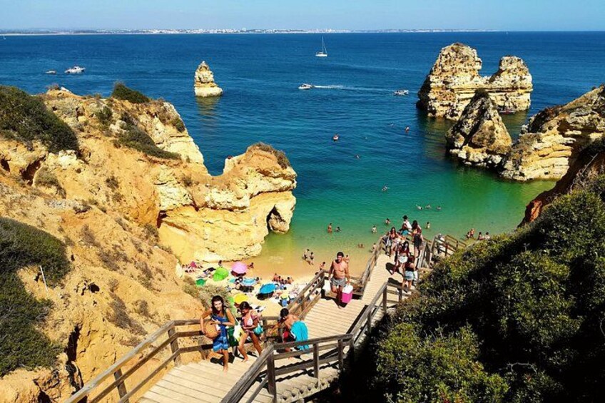 A coastal area with a wooden walkway leading to a beach, rocky cliffs, and clear blue water. 1