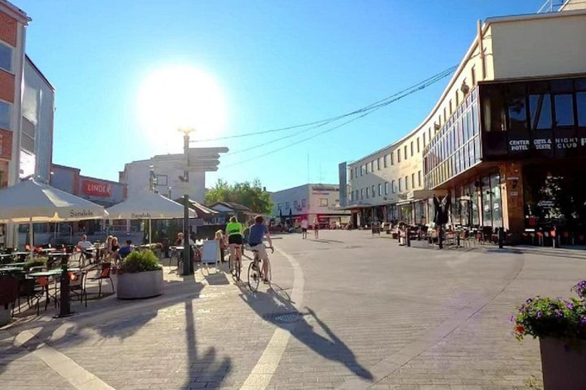 A sunny urban square with outdoor seating, a cyclist, and buildings with commercial signs. 2