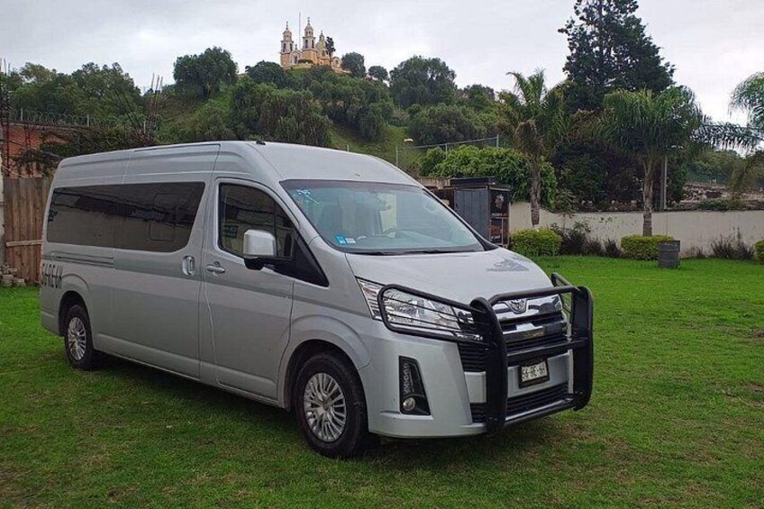 A silver minivan parked on a grassy area with a backdrop of trees and a distant building. 3