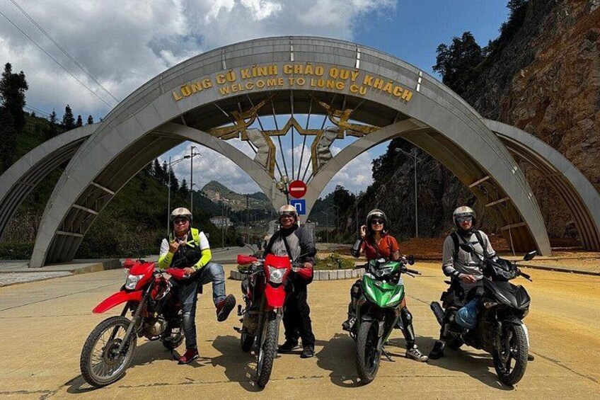 Three motorcyclists in front of a large archway with text welcoming to Lung Cuu Khach. 1