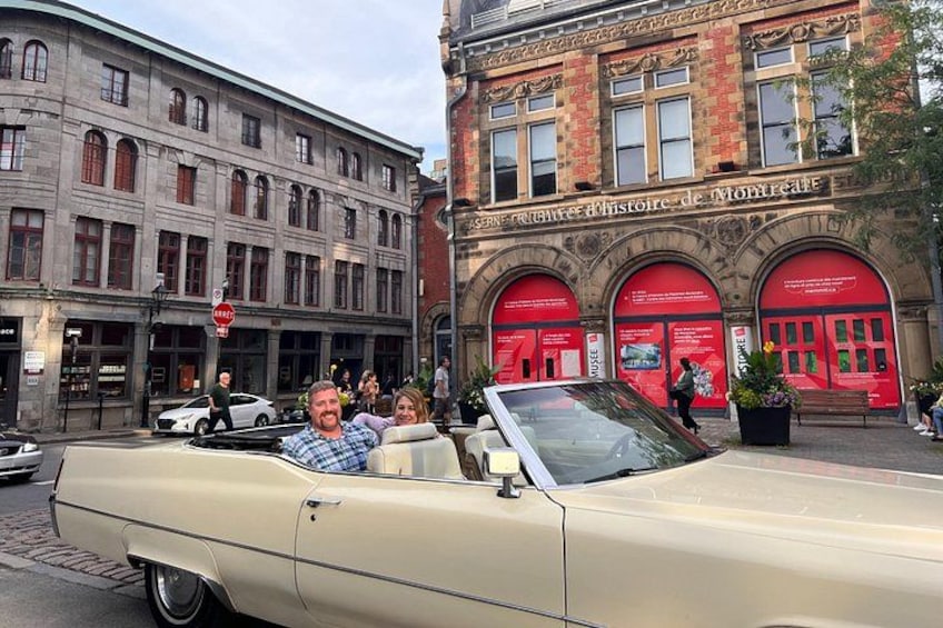 A beige convertible car parked on a street with a red building in the background. 4