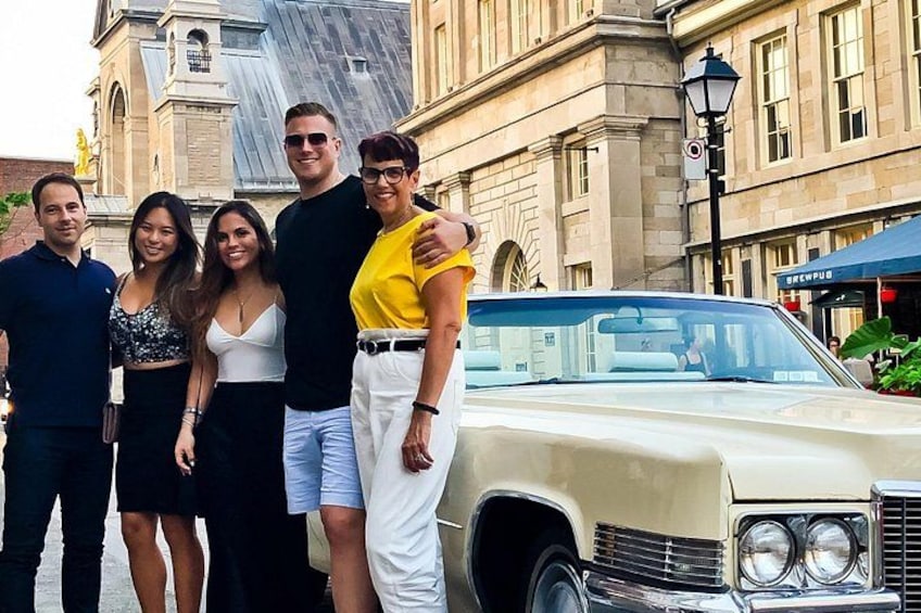 A group of people posing in front of a vintage car with a building in the background. 1