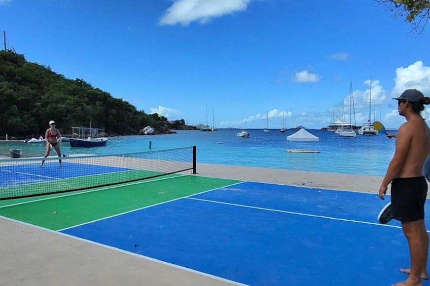 Cancha de tenis de playa con red, dos jugadores y un fondo costero con barcos y cielo despejado. 4