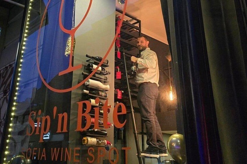 Una persona su una scala all'interno di un edificio, probabilmente un ristorante, con un cartello che riporta 'Sip'n Bite' e 'Sofia Wine Spot' visibile attraverso una finestra. 2