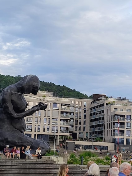 Suuri veistos, jossa on kuvattu istuva hahmo kaupunkimaisemassa, taustalla moderneja rakennuksia ja rinne. 2