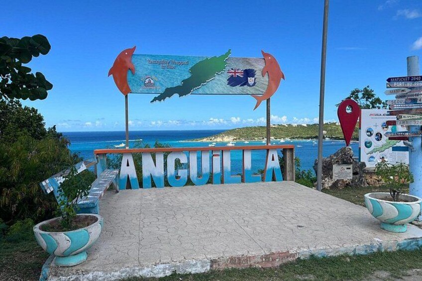 Un cartel con la palabra 'ANGUILLA' y un mapa, rodeado de plantas en macetas y vistas al mar. 1