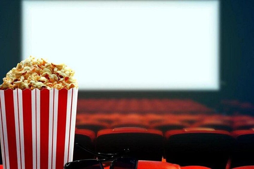A bucket of popcorn in a red and white striped container, a pair of glasses, and a movie screen in a dimly lit theater. 4