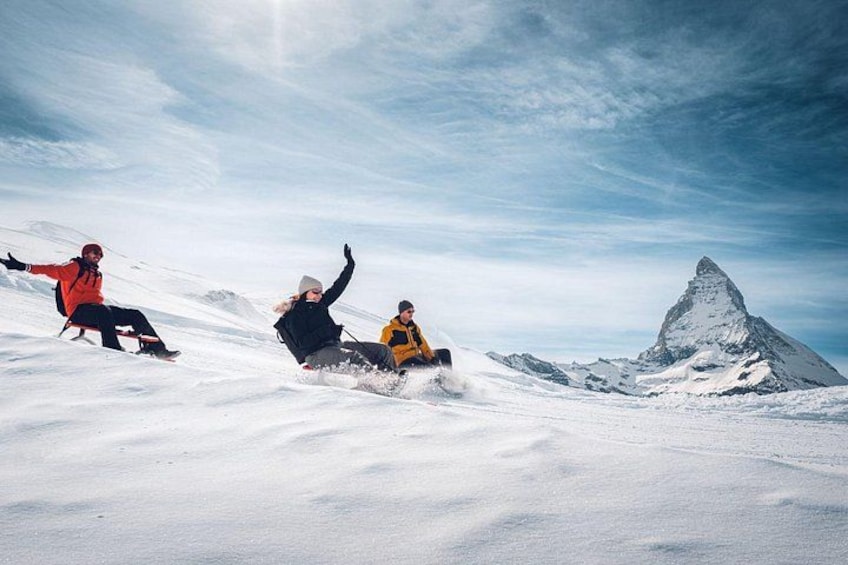 Drie mensen glijden op sledes een besneeuwde helling af, met een bergtop op de achtergrond. 3
