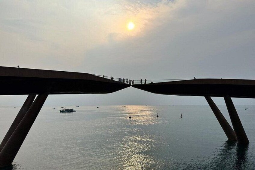 A bridge with people walking on it, overlooking a calm body of water with boats and a setting sun. 2