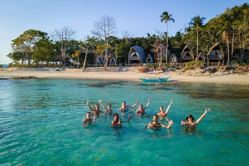 People swimming in clear blue water with a beach and huts in the background. 1