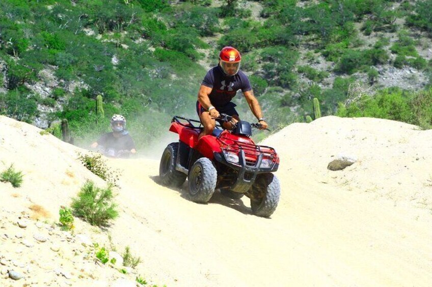 Een persoon rijdt op een rode quad op een onverhard pad, met schaarse begroeiing en een rotsachtige heuvel in de achtergrond. 2