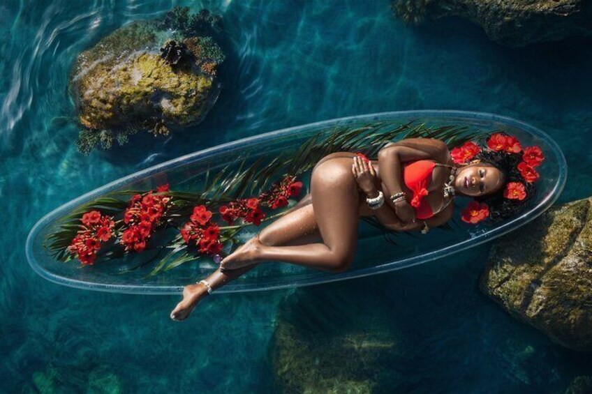 A person floating on a clear oval object in a body of water surrounded by rocks and red flowers. 1