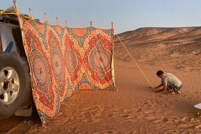 A person in a desert setting, a colorful patterned cloth hanging between two poles, and a vehicle with a spare tire. 5