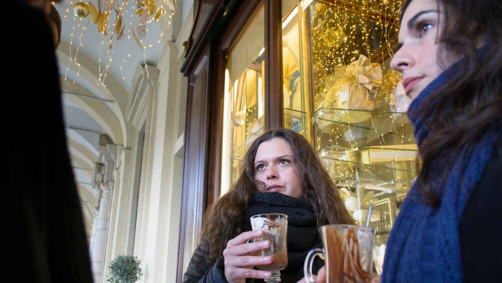 Two people holding mugs, possibly with drinks, in front of a building with ornate architecture and string lights. 5