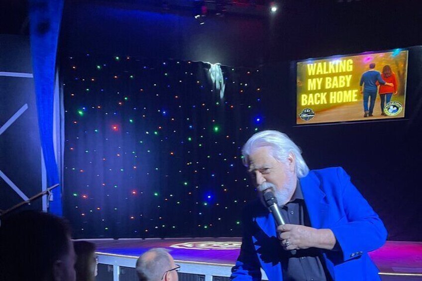 A person in a blue suit speaking into a microphone on a stage with a backdrop of colorful lights and a sign reading "Walking My Baby Back Home". 2