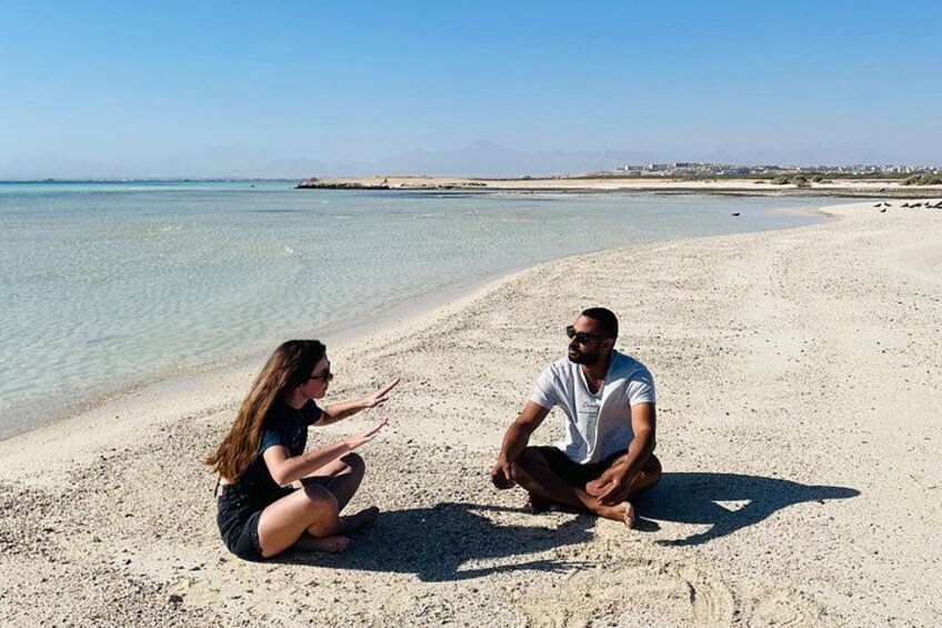 Dos personas sentadas en una playa arenosa con cielo despejado y mar en calma al fondo. 2